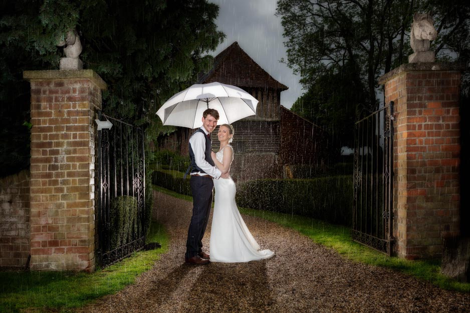 Bride and groom under an umbrella in front of the brick pillars at Colville Hall